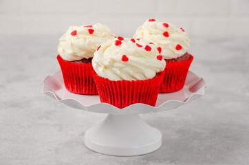 Red velvet cupcakes with cream cheese icing are decorated for Valentine's Day on a white stand on a gray background. Copy space.