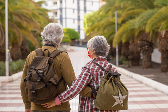 Back View Of Two White-haired Traveler People Walking Together During The City Tour, Wearing A Surgical Mask Due To The Coronavirus Active Retired People Enjoying Travel And Freedom