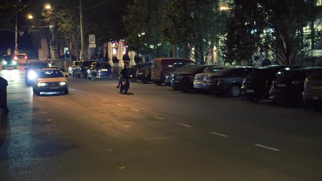 Attractive rider man starting from parking place on motorbike, turns on and riding by night city street