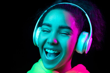 Fototapeta premium Laughting, close up. African-american woman isolated on dark background in multicolored neon. Listening to music with headphones. Concept of human emotions, facial expression, sales, ad, fashion.
