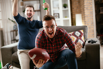 Two young friends enjoying at home. Men drinking beer and watching sports game on tv