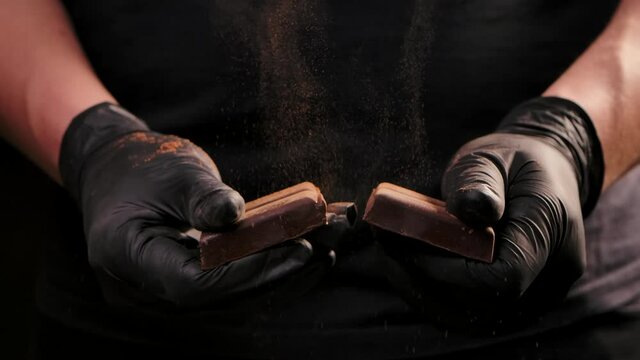 Man hands in black gloves breaking chocolate. Dark chocolate bar breaking in half. Sweet ingredient for cooking dessert. Close up. Slow motion.