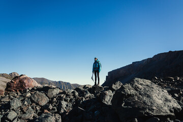 Young woman traveler in the mountains with a backpack with trekking poles stands on a large stone against a cloudless blue sky. Social distancing concept when traveling
