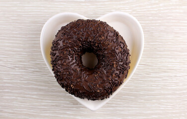 Round sweet donut in chocolate in a plate in the shape of a heart on a light wood background