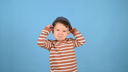 Cute Caucasian little boy standing against blue background making grumpy face. Funny emotional child makes a grimace and looking at kamera. Boy in hat and striped swetear