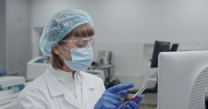 Close up of female medical worker in protective mask and googles entering data on computer. Scientist in rob and bouffant cap holding glass test sample tube while typing in laboratory.
