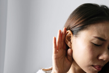 Fototapeta premium Closeup of Asian girl holds her hand near ear and listens carefully on grey background. Hearing problem, deaf concept.
