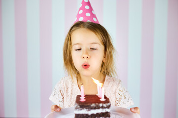 Little girl in pink cap blowing out candles on a birthday chocolate cake