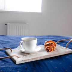 Classical breakfast. A cup of coffee and a croissant stand on a white wooden tray that stands on the bed