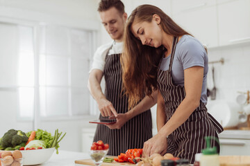 Happy couple love cooking together in kitchen. Young loving man and woman cooking and watching online cookbook in white kitchen room.