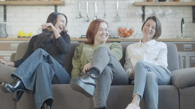 Three cheerful women crossing legs and laughing sitting on couch with kitchen at background. Positive Caucasian female friends having fun posing indoors at home. Friendship and leisure.
