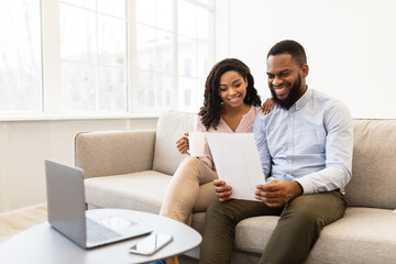 Positive black couple reading letter at home, sitting on couch - Powered by Adobe