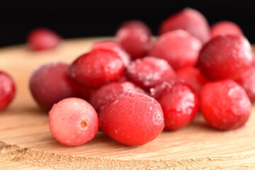 Several berries of sweet and sour freshly frozen cranberries on a round wooden tray, close-up, black background.
