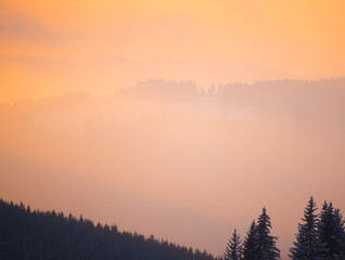 Abstract background of dreamy mauve pink to pastel golden orange fog light over mountain forest silhouettes at winter sunrise or sunset. Magic Valentine moments in foggy mountain valley landscape. 