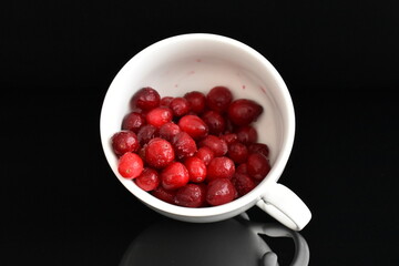 Several berries of natural freshly frozen cranberries in a white ceramic cup, close-up, black background.