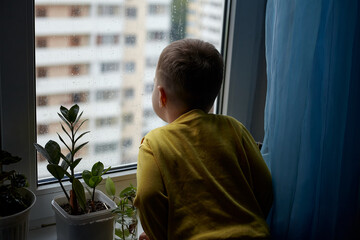 A little boy looks sadly out of the window. The concept of rainy weather. The child stays at home because of the rain