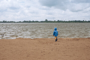 child running on the beach. boy in blue jacket and rain boots stands in front of a large sea on the beach. On the beach in autumn. With a jacket on a windy beach