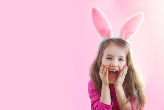 A little cheerful girl in rabbit ears opened her mouth wide and holds her cheeks with her hands on a pink background. Banner. Happy Easter holiday concept. Copyspace
