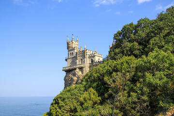 Fototapeta na wymiar View of the Swallow's nest from the shore on a sunny day. Bright blue sky on a summer day. A large green bush covers the rock with a castle.