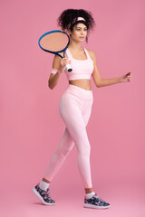 full length of young curly sportswoman holding tennis racket while walking on pink