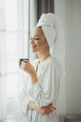 Young woman in bathrobe drinking coffee and looking through the window.