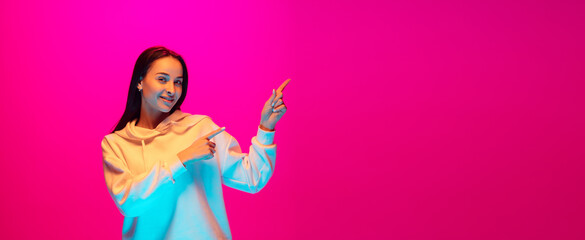 Pointing, flyer. Caucasian woman's portrait isolated on pink studio background in mixed neon light. Beautiful female model. Concept of human emotions, facial expression, sales, ad, fashion. Copyspace.