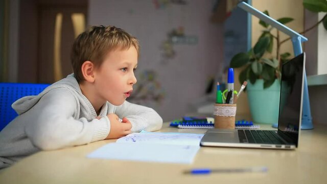 Cute Little Boy Studying At Home With Laptop, Repeating Words After Teacher. Training Books And Notebook On The Table. Distance Learning Online Education