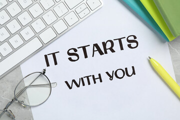 Sheet of paper with phrase It Starts With You, keyboard and stationery on grey table, flat lay
