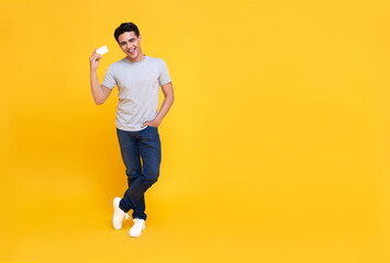 Happy handsome Asian shopaholic man showing credit card in hand isolated on yellow background.