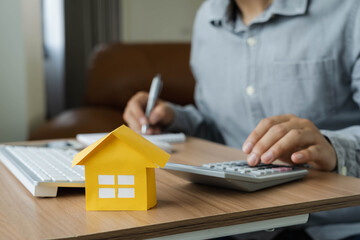 Business  Finance concept. Close up yellow paper house model with young Asian man wear grey shirt ongoing to calculate home loan mortgage to summary expense payment  with using laptop on office deck