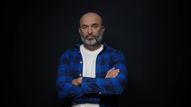 Bearded middle aged man looks disappointed folding one's arms standing over black background. Human emotions.