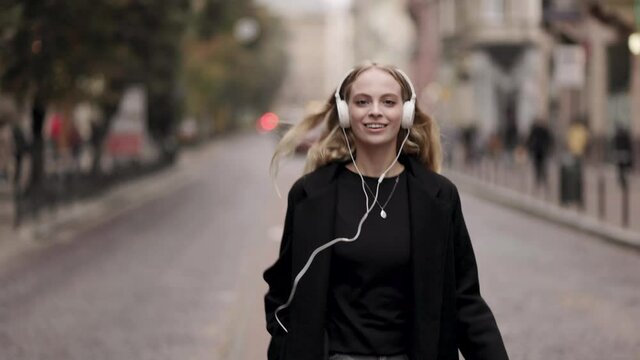 Happy young woman in headphones dancing outdoors in city street having fun alone. Joyful attractive blonde carefree woman listening to music with smartphone