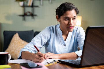 Young business woman writing down notes by looking laptop - concept of employee or student online...