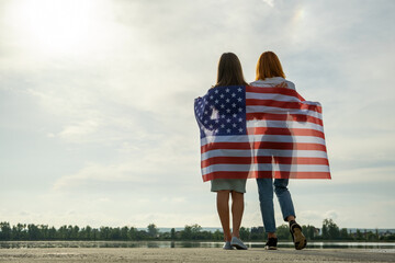 Two young friends women with USA national flag on their shoulders standing together outdoors on...