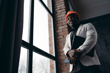 A businessman in an orange protective helmet in a white jacket stands on a light background