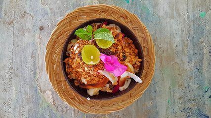 Exotic breakfast bowl indonesian style with yoghurt, granola, fruits, coconut, lime, peppermint with a pink blossom on wooden table in Bali Asia