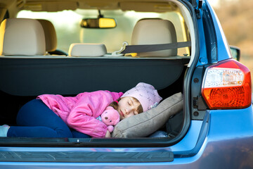 Pretty happy child girl sleeping with a pink toy teddy bear in a car trunk.