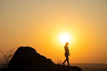 Silhouette of a woman hiker walking down a big stone at sunset in mountains. Female tourist on high rock in evening nature. Tourism, traveling and healthy lifestyle concept.