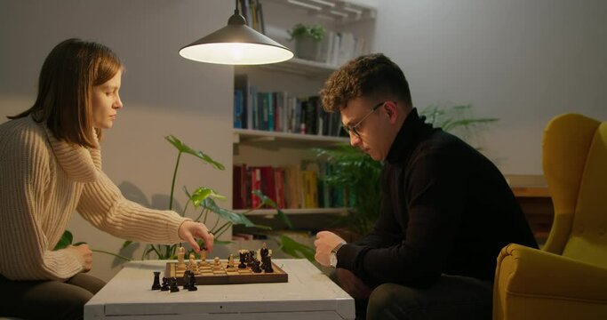 Young Couple Play Chess at Home at Night. Lady wins game. Sport and Hobby Activities at quarantine or isolation at Pandemic. Friends move chess pieces on chessboard with checkmate. 4K Medium Pan Shot