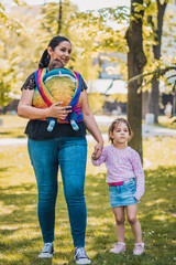 Fototapeta premium Beautiful happy young mother with baby and child carrier in park. Mom walking with infant in baby carrier. Happy mother carrying her child by ergonomic baby carrier.