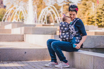 Beautiful happy young mother with baby carrier in park. Mom walking with infant in baby carrier....