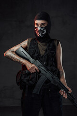 Tattooed and seductive woman in black armour and weared with mask poses in dark background holding assault rifle.