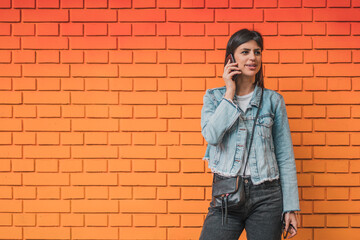 Fototapeta na wymiar Attractive young woman posing and talking on mobile phone on a background of a red colorful brick wall. Empty space for text o design.