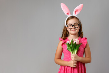 Obraz na płótnie Canvas Cute happy little girl wearing easter bunny ears and glasses on gray background. The child is holding a bouquet of tulips.