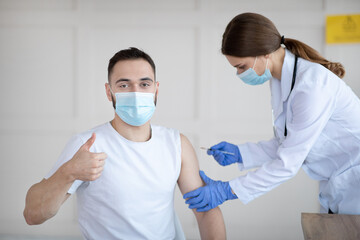 Fototapeta na wymiar Millennial man in mask recommending vaccination against covid-19, showing thumb up gesture while getting vaccine shot