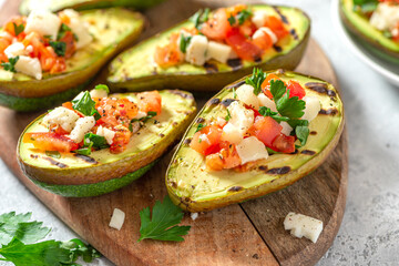 Grilled avocado stuffed with tomatoes, goat cheese, parsley, and olive oil on a cutting board close-up.