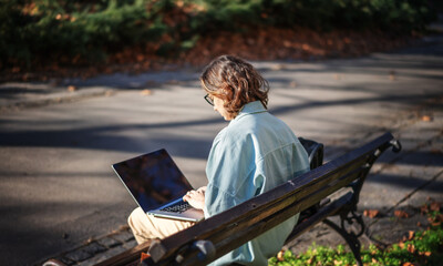 A young serious woman student sitting on a bench in the park and typing on her laptop on a sunny day in universiry campus