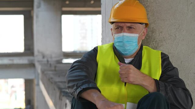 Builder in medical protective face mask and hard hat sits at construction site. He coughs and he is not feeling well, he has chest pain