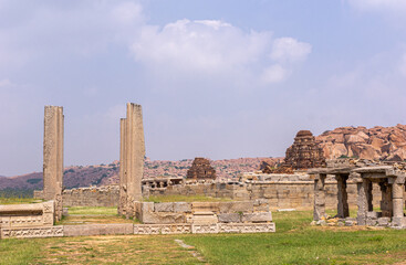 Fototapeta premium Hampi, Karnataka, India - November 5, 2013: Vijaya Vitthala Temple red stone gopurams behind beige stone ruins of ancient temeple under light blue cloudscape, Grass up front.