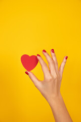 Woman hand with red fingernails holds a paper heart on yellow background. Love sign, Valentines day concept.